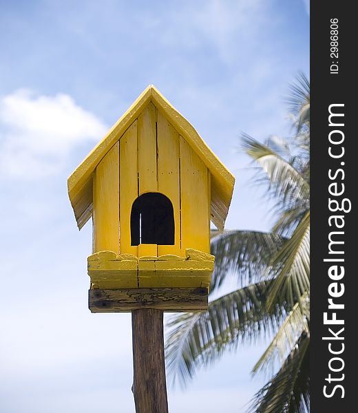 Yellow birdhouse on a background of palm trees (out of focus) and cloudy, blue sky.