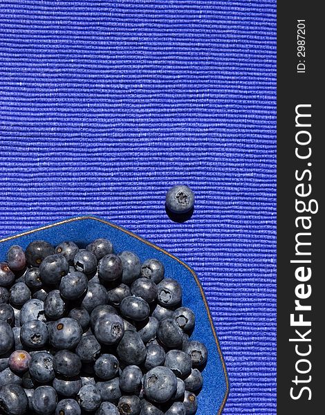 A blue theme for summer fruit -- a single berry has escaped from a blueberry-filled blue bowl on a blue placemat. A blue theme for summer fruit -- a single berry has escaped from a blueberry-filled blue bowl on a blue placemat.