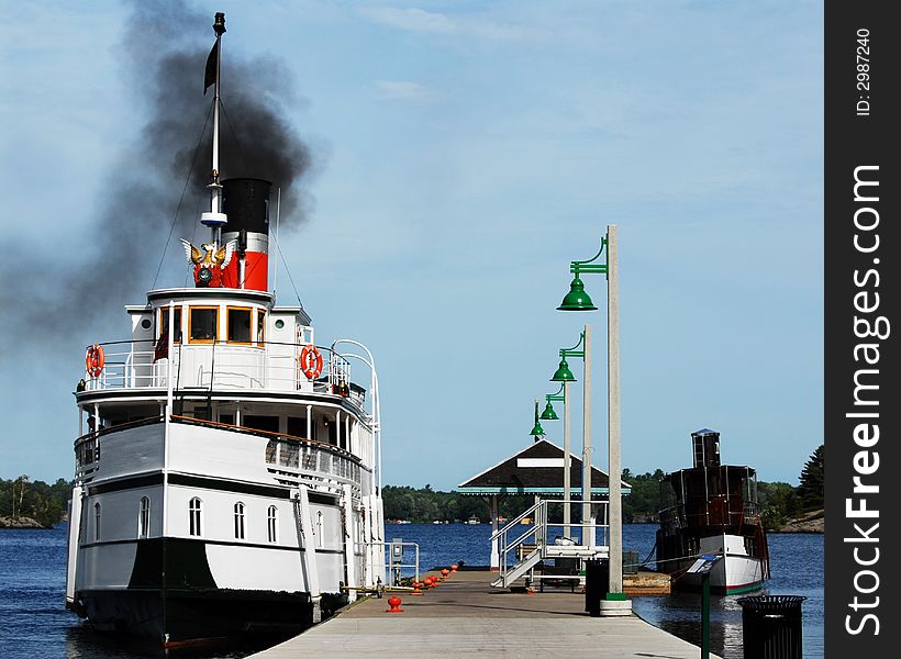 Two vintage steamships at dock.  The larger, white one is puffing dark black smoke.