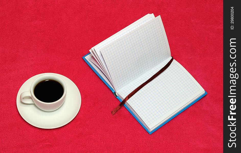 The cup of coffee and open notebook on a red blanket. The cup of coffee and open notebook on a red blanket