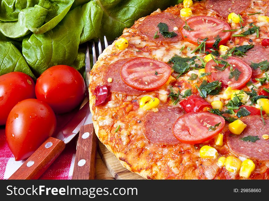 Salami pizza sitting on wooden cutting board with tomatoes and salad