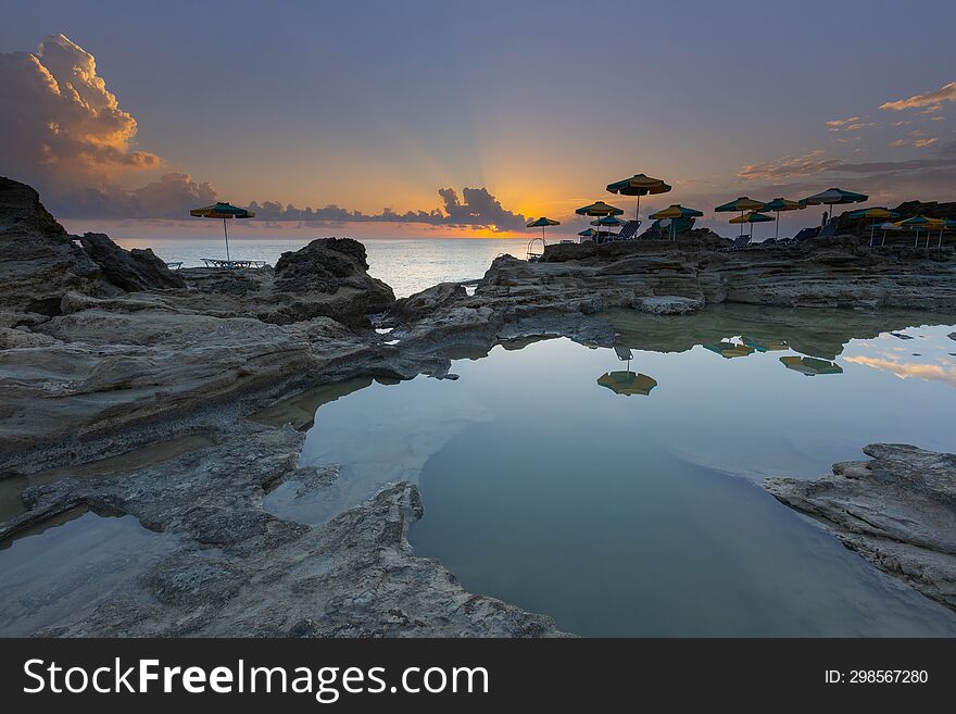 A romantic bay by the sea at sunrise with small lagoons in which the sky is reflected. Among the lagoons, there are umbrellas with sunbeds on the stones