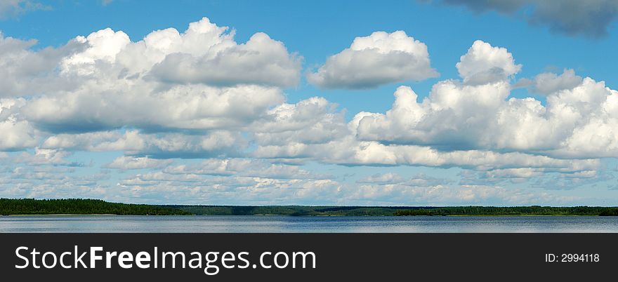 White fluffy clouds, blue sky and green forest on far bank of the lake.