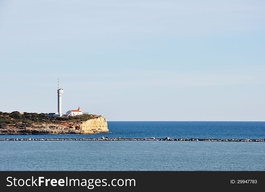 Image shows lighthouse of Portimao, Algarve, Portugal, Europe. Lighthouse is on the left, sea in front, clear blue sky in the back.