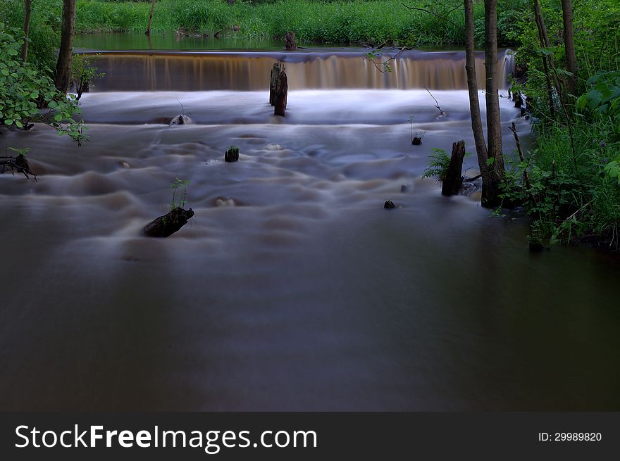 The photo shows a small waterfall on a small river. It is located in the forest, surrounded by greenery. The photo shows a small waterfall on a small river. It is located in the forest, surrounded by greenery.