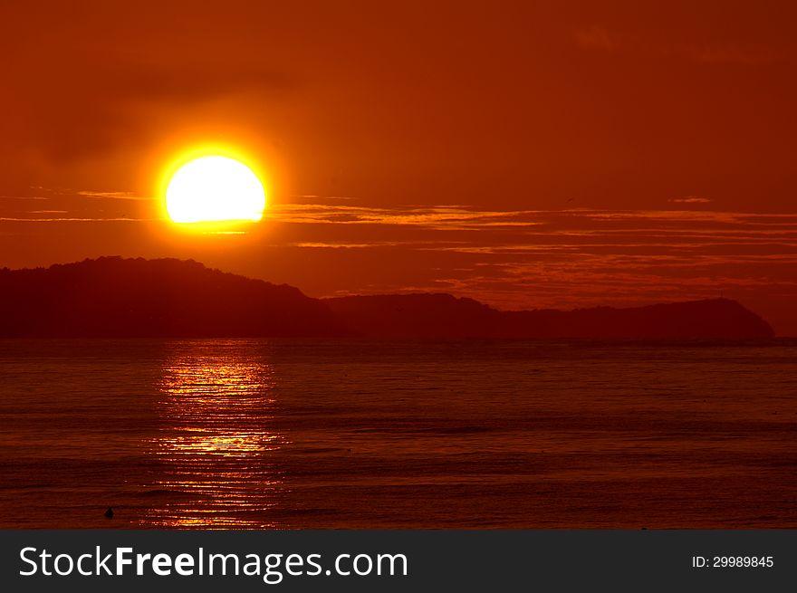 The photo shows the shield of the sun at high zoom settings. The sun is low on the horizon. It is a small part hidden behind the clouds. Sunlight reflected from the surface is located in the foreground of the sea. The photo shows the shield of the sun at high zoom settings. The sun is low on the horizon. It is a small part hidden behind the clouds. Sunlight reflected from the surface is located in the foreground of the sea.