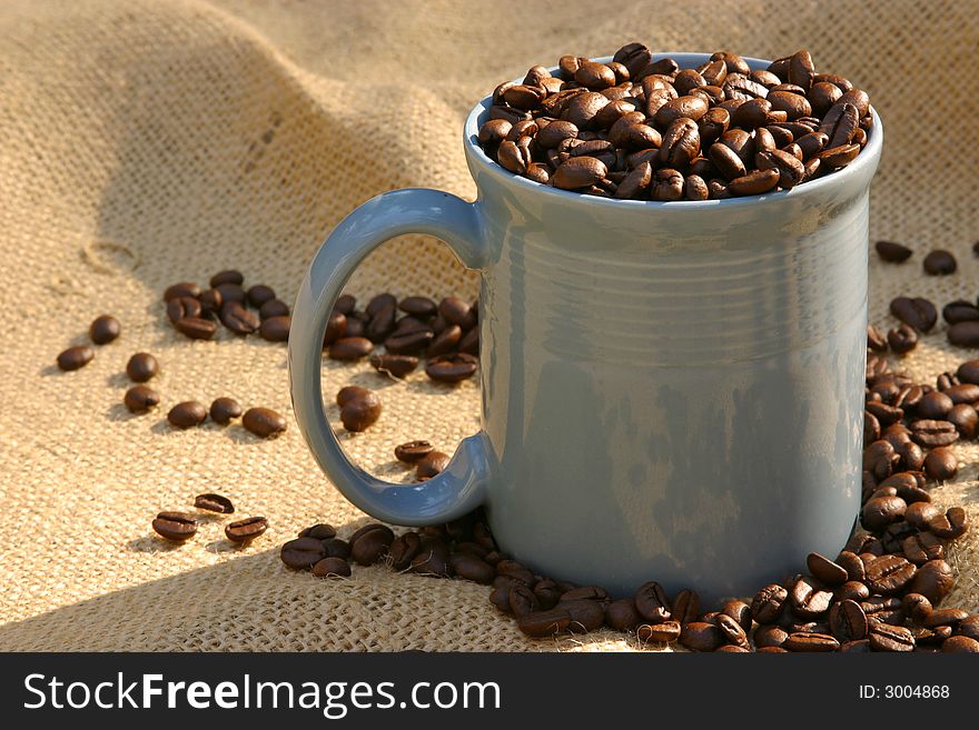Blue mug of roasted coffee beans on burlap in morning sun. Blue mug of roasted coffee beans on burlap in morning sun.