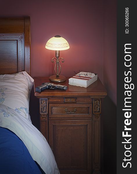 Glock on night table.With a night lamp, book and glasses beside a blu bed. This is ready to read a thriller book maybe and he's afraid to be scared