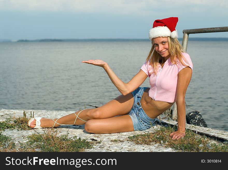 The young beautiful woman sits on seacoast with Santa Claus hat. The young beautiful woman sits on seacoast with Santa Claus hat