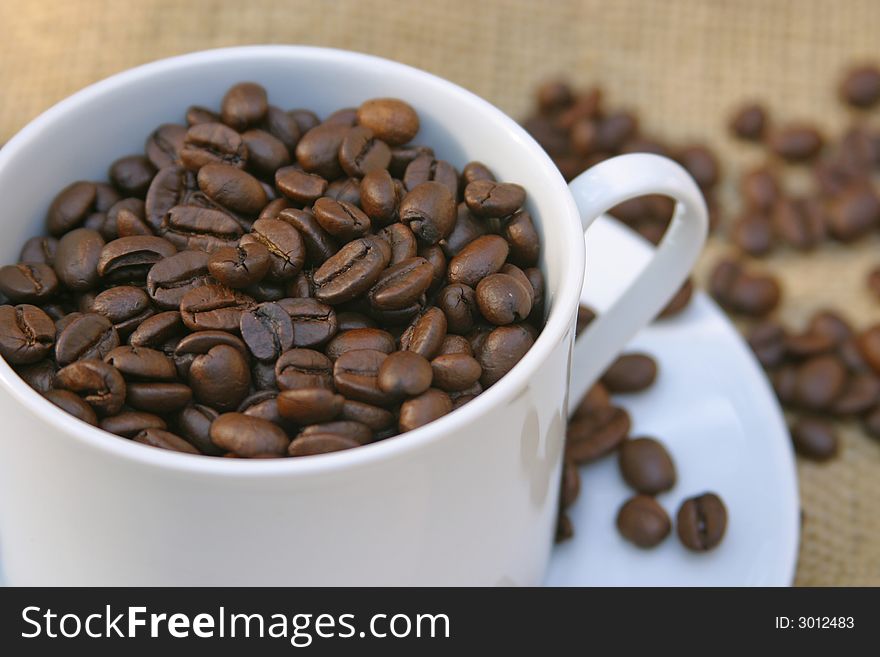 White cup of roasted dried coffee beans on burlap. White cup of roasted dried coffee beans on burlap.