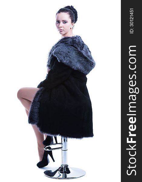 Portrait of beautiful face brunette model sitting on the bar chair with high-heel shoes, wearing luxury black color fur coat, looking back over shoulder straight to the camera. Isolated on white background. Shot in studio. Portrait of beautiful face brunette model sitting on the bar chair with high-heel shoes, wearing luxury black color fur coat, looking back over shoulder straight to the camera. Isolated on white background. Shot in studio.