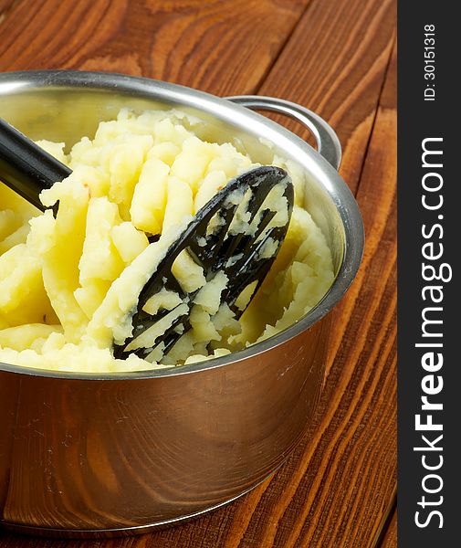 Preparing Mashed Potato in Stainless Steel Pan with Black Potato Masher on Wooden background