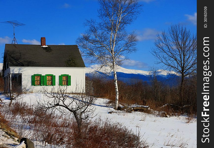 Abandoned house with green shutters in snow with blue sky and birch trees and White Mountains in background. Abandoned house with green shutters in snow with blue sky and birch trees and White Mountains in background