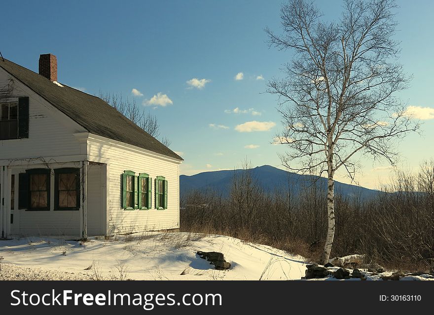 Abandoned house with green shutters in snow with blue sky and birch tree and mountain in background. Abandoned house with green shutters in snow with blue sky and birch tree and mountain in background