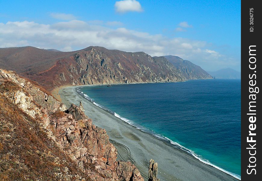 A cloudscape on sea: turquoise sea, grey beach, yellow-red capes and rocks, white surf and white clouds on azure sky. Autumn. Russian Far East, Primorye, Japanese sea, Tasovaya bay. A cloudscape on sea: turquoise sea, grey beach, yellow-red capes and rocks, white surf and white clouds on azure sky. Autumn. Russian Far East, Primorye, Japanese sea, Tasovaya bay.