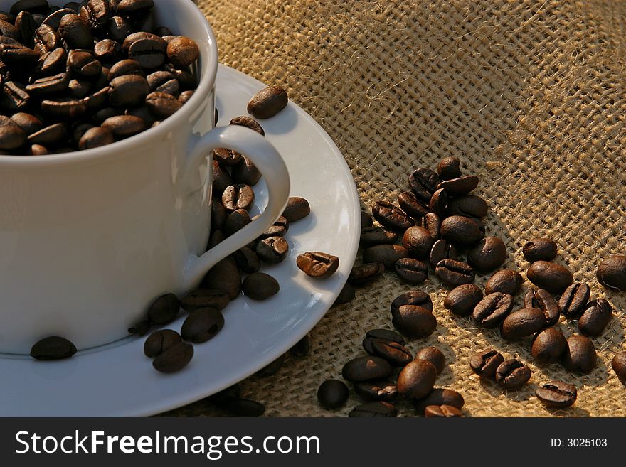 White cup and saucer filled with roasted dried coffee beans on burlap in early daylight. White cup and saucer filled with roasted dried coffee beans on burlap in early daylight..