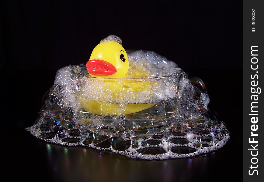 Bright yellow rubber duck taking a bubble bath in a small, clear glass dish with bubbles falling all around the outside of the dish.  This photo has a black background. Bright yellow rubber duck taking a bubble bath in a small, clear glass dish with bubbles falling all around the outside of the dish.  This photo has a black background.