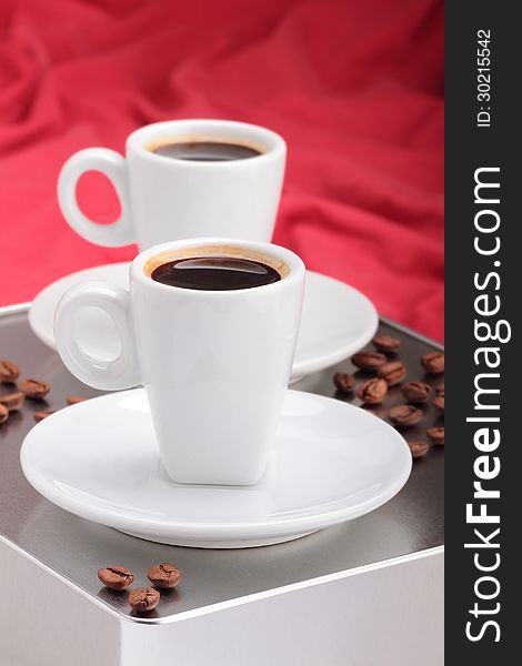 Two espresso cups on a gray metallic table, on red background blur