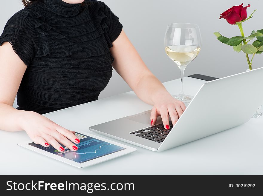 A business woman is working with the modern laptop and tablet computer. Nearby on the table is a glass of wine and one rose. A business woman is working with the modern laptop and tablet computer. Nearby on the table is a glass of wine and one rose.