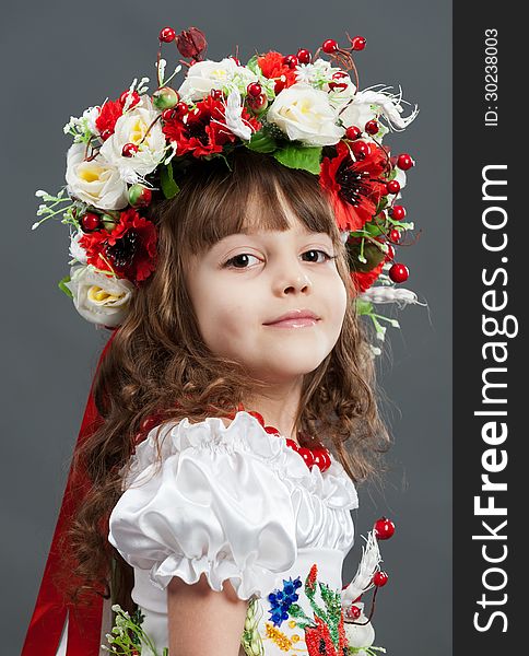 Close-up portrait of cute little girl dressed in folk costumes with large bright wreath on her head on gray background in the studio. Close-up portrait of cute little girl dressed in folk costumes with large bright wreath on her head on gray background in the studio