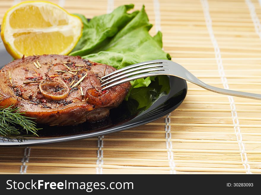 Grilled steak with salad and lemon on black plate with fork. Bamboo background