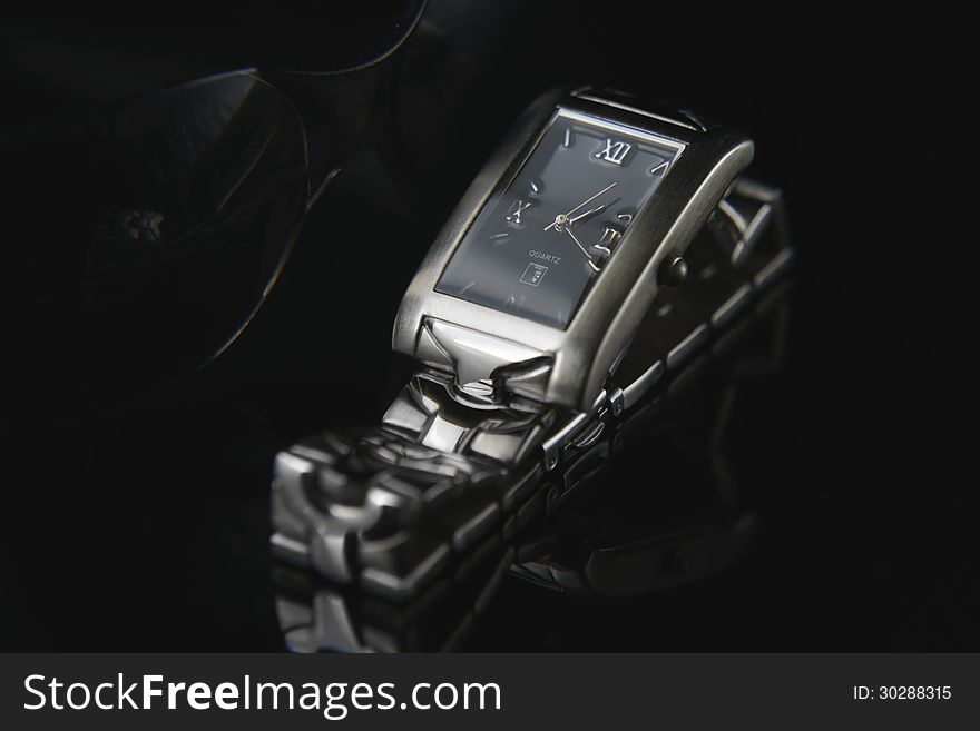 Close up of wrist watch casting reflection on black. Black color background with black sunglasses. Close up of wrist watch casting reflection on black. Black color background with black sunglasses.