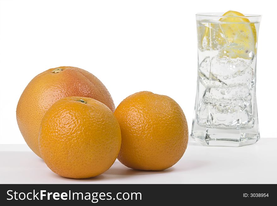 Two oranges and a grapefruit with a glass of sparkling water in the back filled with ice and a slice of lemon. Two oranges and a grapefruit with a glass of sparkling water in the back filled with ice and a slice of lemon