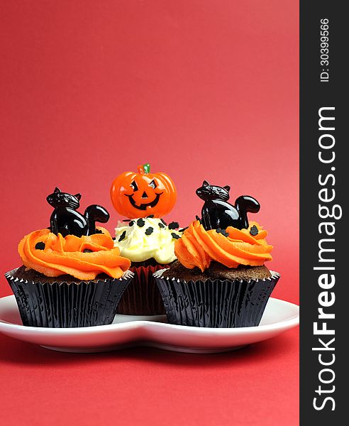 Happy Halloween orange and black decorated cupcakes with black cats and jack-o-lanterns on white heart plate against a red background. Happy Halloween orange and black decorated cupcakes with black cats and jack-o-lanterns on white heart plate against a red background.