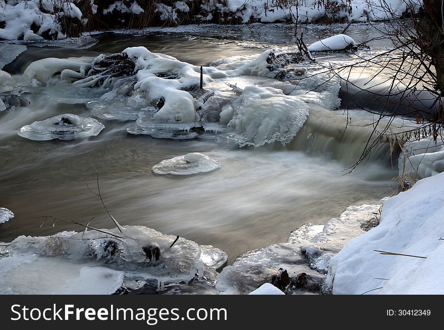 The photograph shows a small waterfall on the river in winter scenery. The banks of the river are covered with a layer of snow. In the water we see the ice caps. The photograph shows a small waterfall on the river in winter scenery. The banks of the river are covered with a layer of snow. In the water we see the ice caps.