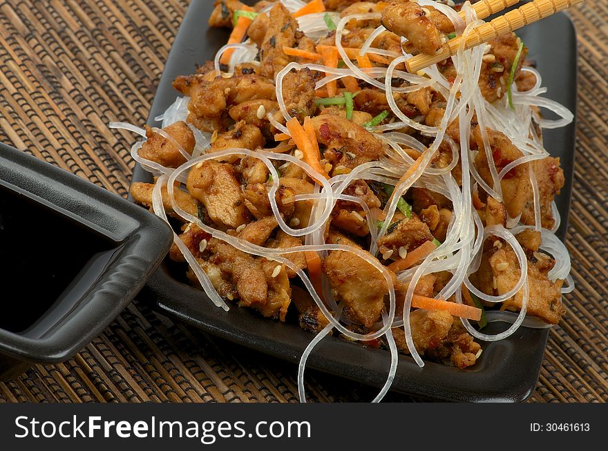 Picked up Piece of Chicken Teriyaki From Bowl by Pair of Chopsticks and Soy Sauce in Black Square Bowl closeup on Straw mat background