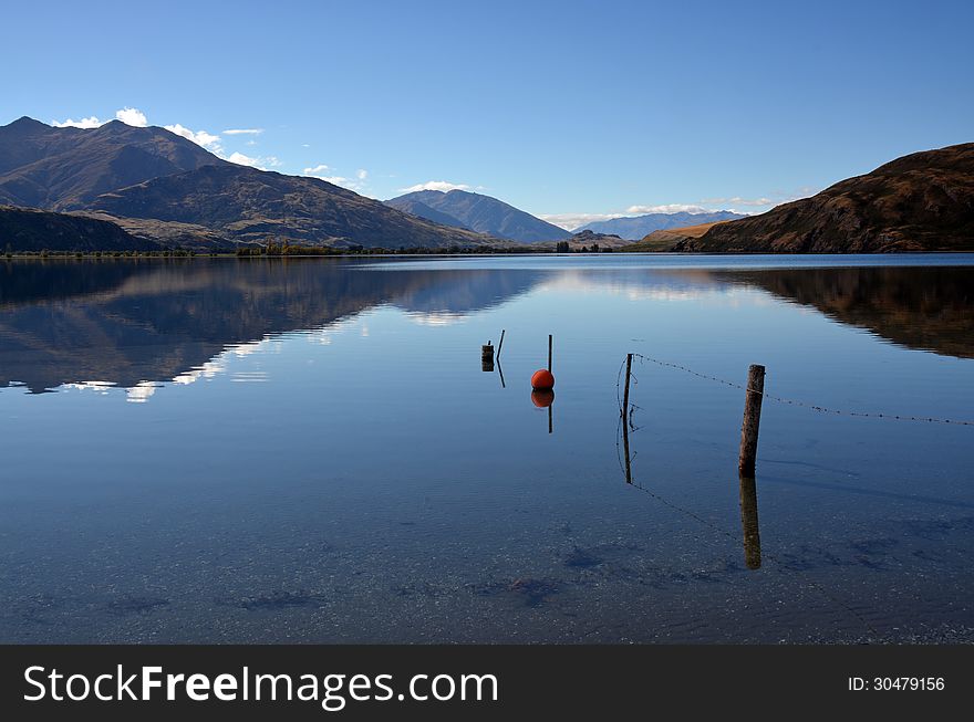 Lake Wanaka reflections in Autumn. In the foreground is a sunken fence and orange buoy. Otago, New Zealand. Lake Wanaka reflections in Autumn. In the foreground is a sunken fence and orange buoy. Otago, New Zealand.