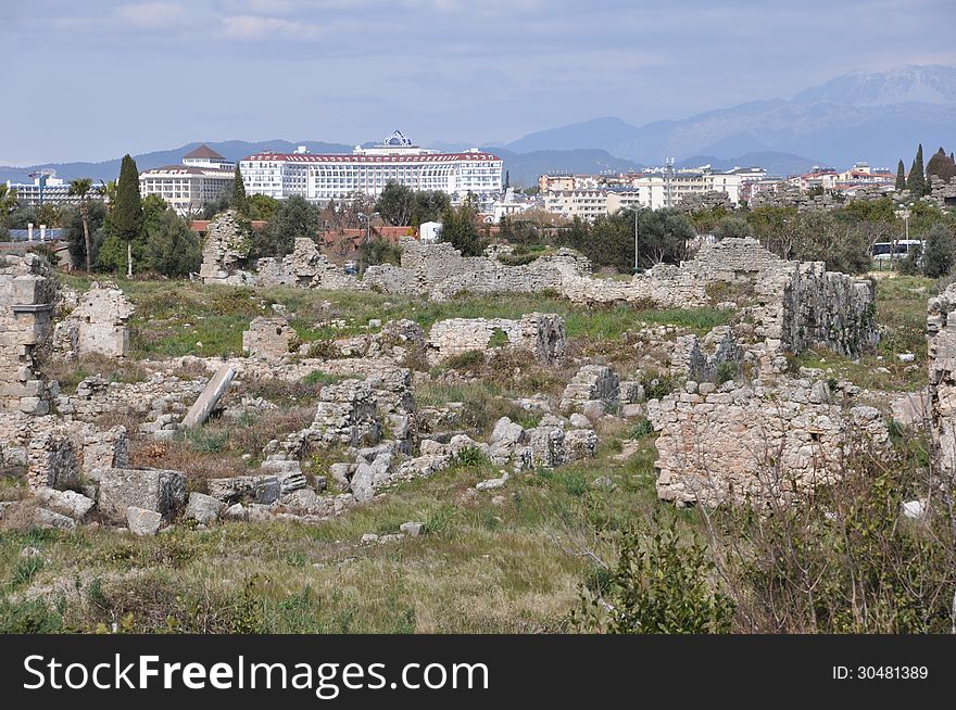 Image shows details of Antique Side, Turkey, with some hotels in the back. Image is dominated by ruins in the front and in the middle. Sky is blue and light cloudy. Image shows details of Antique Side, Turkey, with some hotels in the back. Image is dominated by ruins in the front and in the middle. Sky is blue and light cloudy.