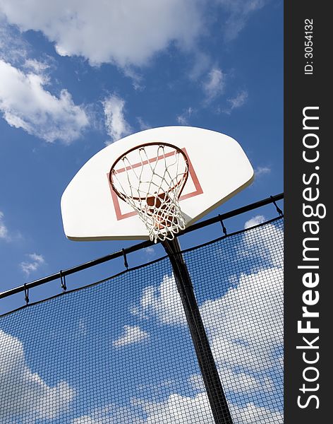 A basketball hoop in a children's playground stands against a partly cloudy blue sky.