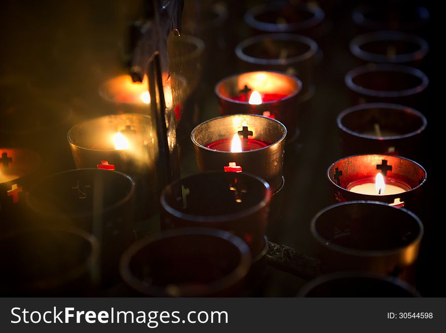 Devotional candles lighting a darkened Roman Catholic Basilica of the National Shrine of Mary, Queen of the Universe. Image shows candle holders, some empty, some full and three with lit candles. Candles are reflected in a gold colored backdrop and 3/4 of a cross is visible in the light. Shot with candle light only. Devotional candles lighting a darkened Roman Catholic Basilica of the National Shrine of Mary, Queen of the Universe. Image shows candle holders, some empty, some full and three with lit candles. Candles are reflected in a gold colored backdrop and 3/4 of a cross is visible in the light. Shot with candle light only.