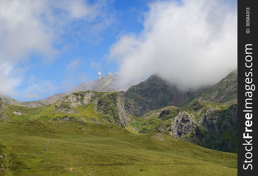 There are green slopes of mountains from the pass of Tourmalet in Pyrenees. In the background the remote peak (Pic du Midi) with an astronomical observatory is covered with white clouds. There are green slopes of mountains from the pass of Tourmalet in Pyrenees. In the background the remote peak (Pic du Midi) with an astronomical observatory is covered with white clouds.