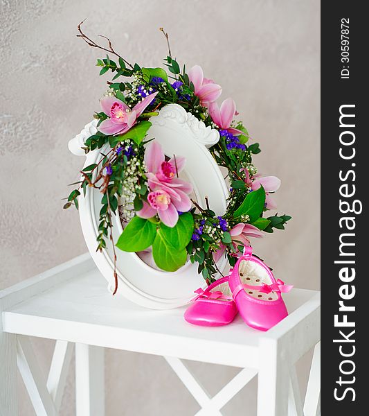 Still life with orchid wreath hanging on the retro style photo frame and infant shoes standing on white wooden stand. Still life with orchid wreath hanging on the retro style photo frame and infant shoes standing on white wooden stand