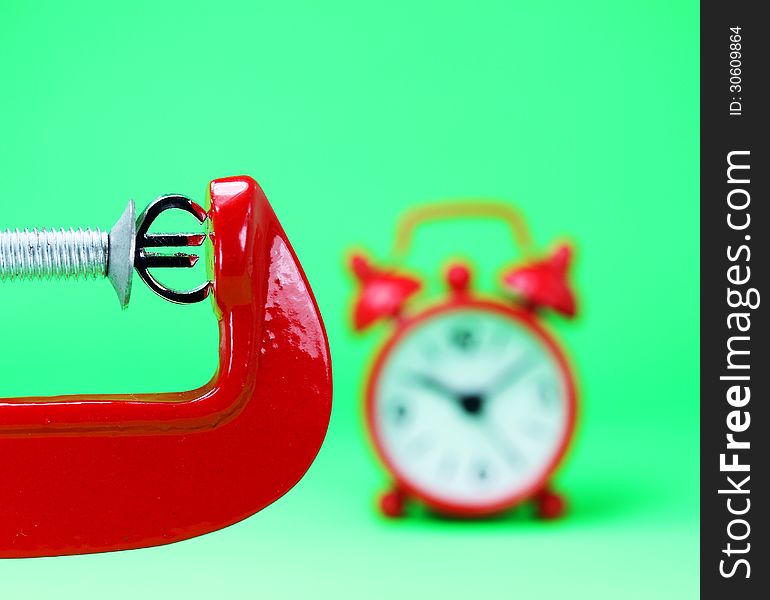 A silver Euro symbol placed in a red clamp with a pastel green background, with a red alarm clock in the background indicating the pressure on the Euro. A silver Euro symbol placed in a red clamp with a pastel green background, with a red alarm clock in the background indicating the pressure on the Euro.