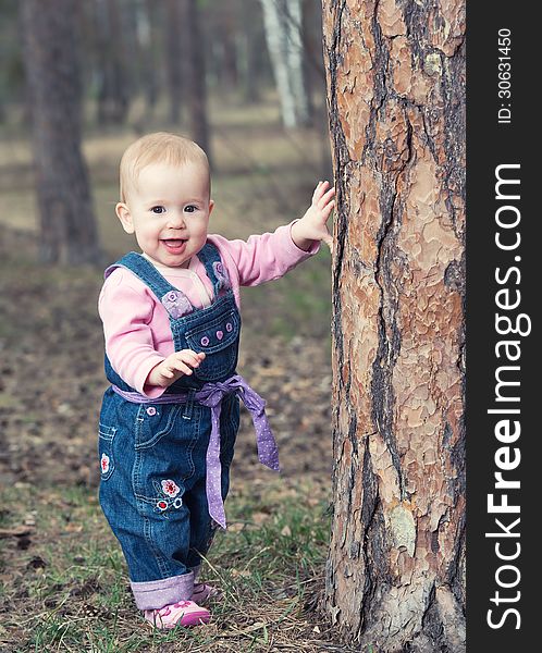 Happy baby girl in jeans jumpsuit stands on legs near a tree in the park outdoors. Happy baby girl in jeans jumpsuit stands on legs near a tree in the park outdoors