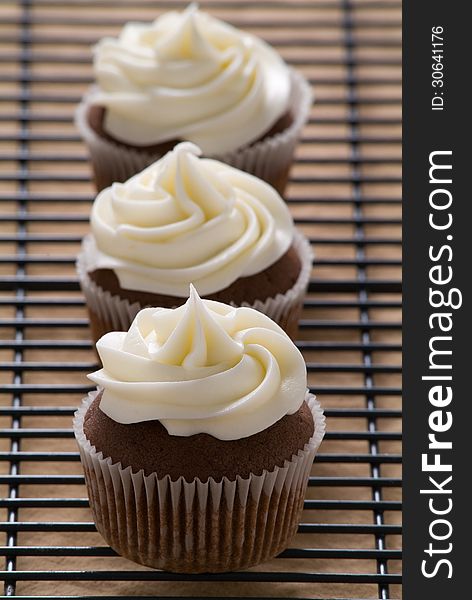 A row of three chocolate cupcakes with vanilla frosting on a cooling rack. A row of three chocolate cupcakes with vanilla frosting on a cooling rack.