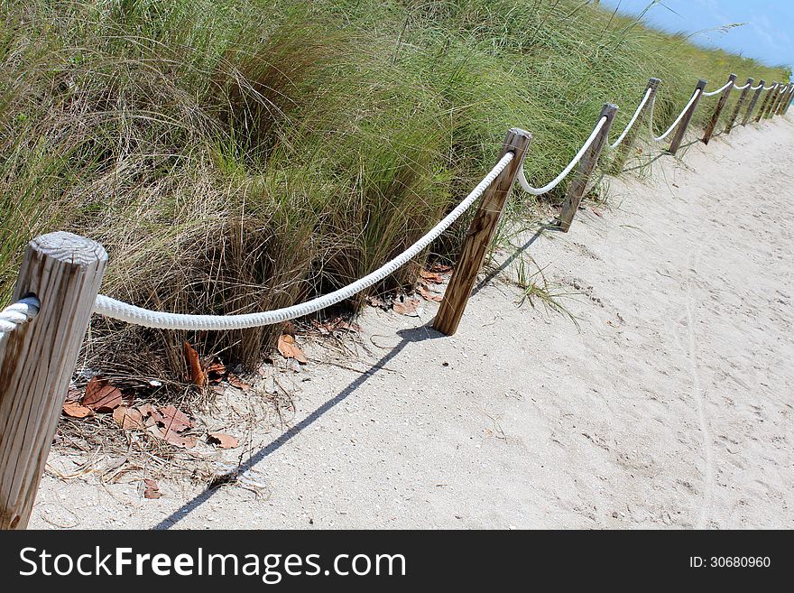Old weathered wood posts with braided ropes running through them, leading a beach-goer through sandy path lined with grasses,onto a warm ocean beach for a leisurely day. Old weathered wood posts with braided ropes running through them, leading a beach-goer through sandy path lined with grasses,onto a warm ocean beach for a leisurely day.