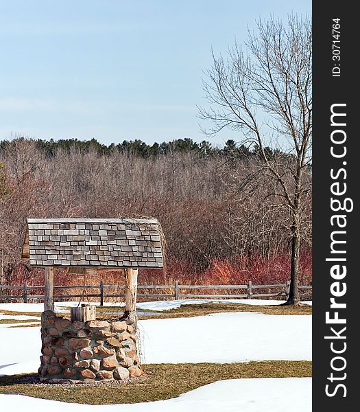 Vertical image of a wishing well in springtime, with snow covering much of the grass.