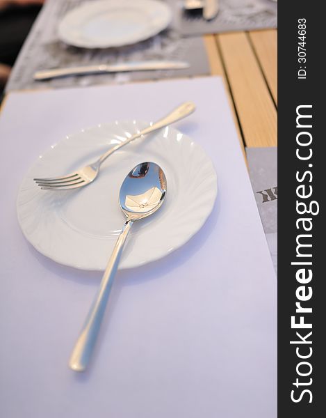 On a white napkin is white plate with a spoon and fork. On a white napkin is white plate with a spoon and fork