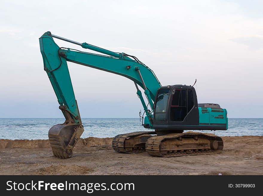 Excavator standing in sandpit with raised bucket at seaside