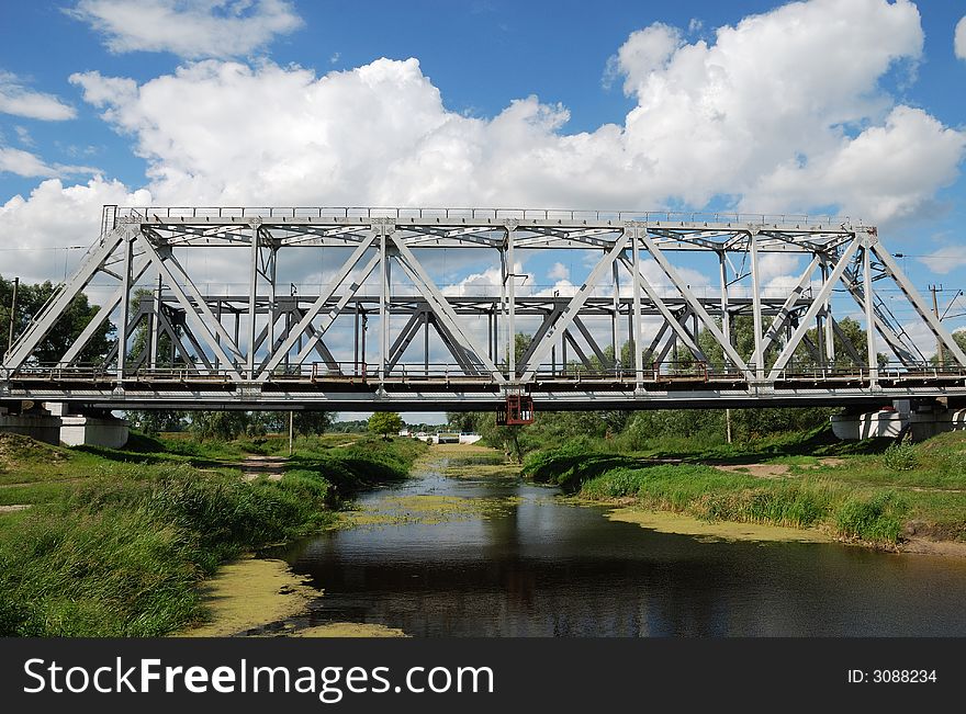 Grey railway bridge over small river with green banks and the cloudy summer sky