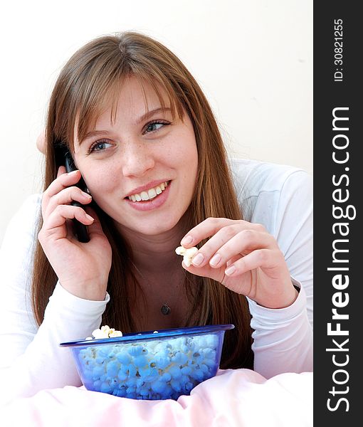 Beautiful young woman enjoying eating popcorn and talking on cell phone on her bed. - Stock Image. Beautiful young woman enjoying eating popcorn and talking on cell phone on her bed. - Stock Image