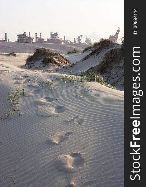 A steel plant with a sandy dune in the foreground. Footsteps in the sand. A steel plant with a sandy dune in the foreground. Footsteps in the sand