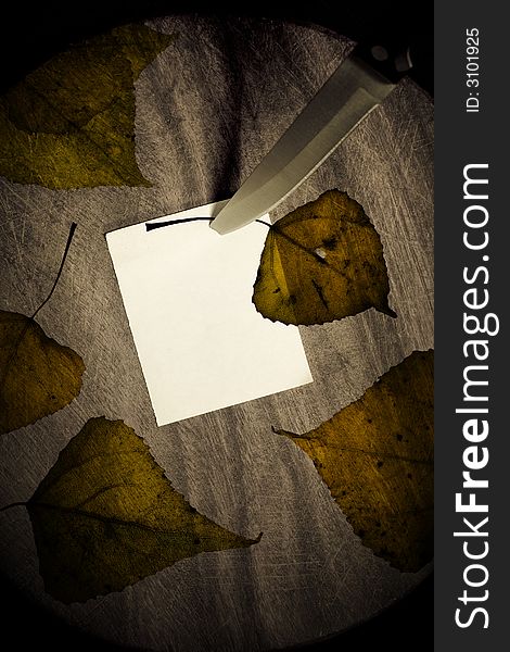 A piece of paper pinned by kitchen knife on  wooden background. Note surrounded by dark brown autumn leaves. A piece of paper pinned by kitchen knife on  wooden background. Note surrounded by dark brown autumn leaves
