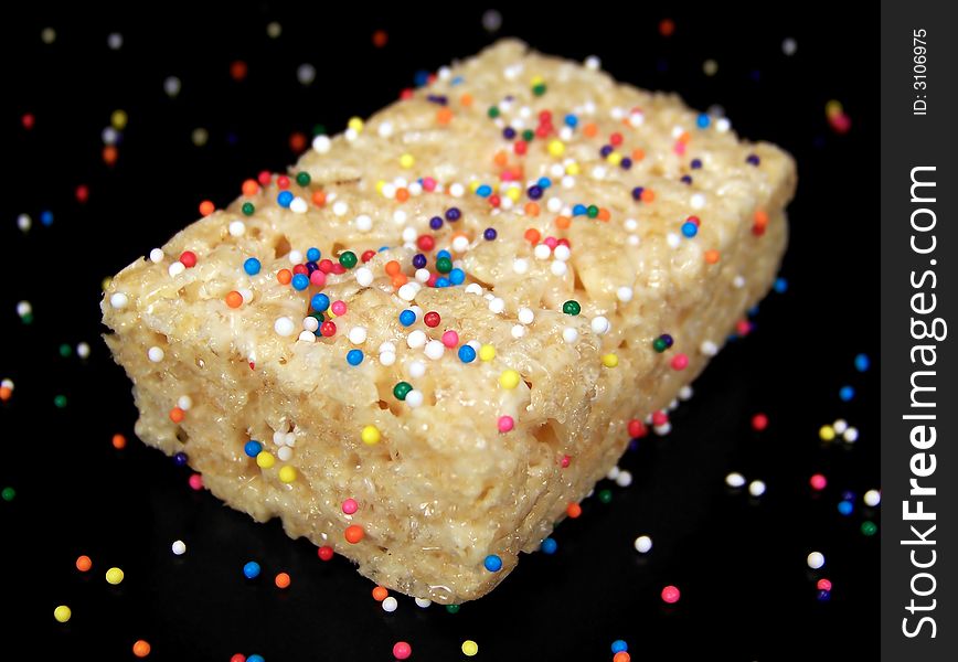 Close up color photograph of a delicious marshmallow crispy dessert bar with colorful sprinkles on and around it.  Black background.