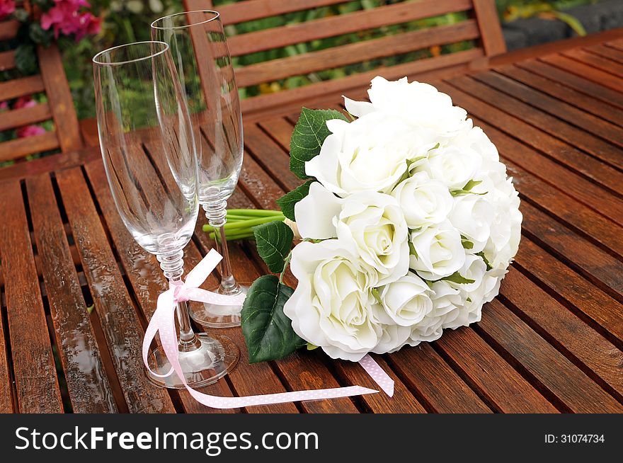 Wedding bridal bouquet of white roses with two champagne glasses with pink polka dot ribbon on outdoor garden table setting after rain.