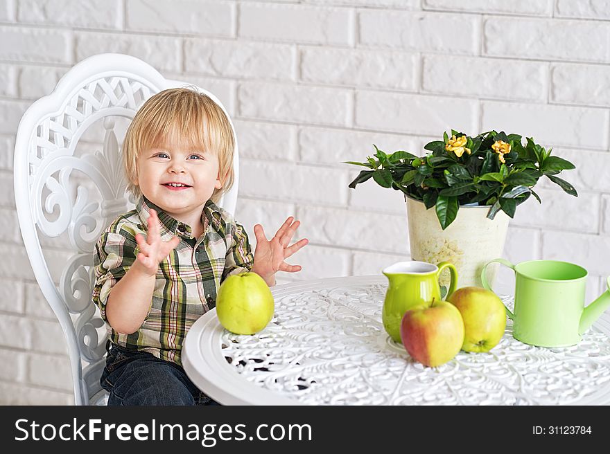 Cheerful baby boy sitting at the vintage style table with pot flower, apples, watering can and a jar. Cheerful baby boy sitting at the vintage style table with pot flower, apples, watering can and a jar